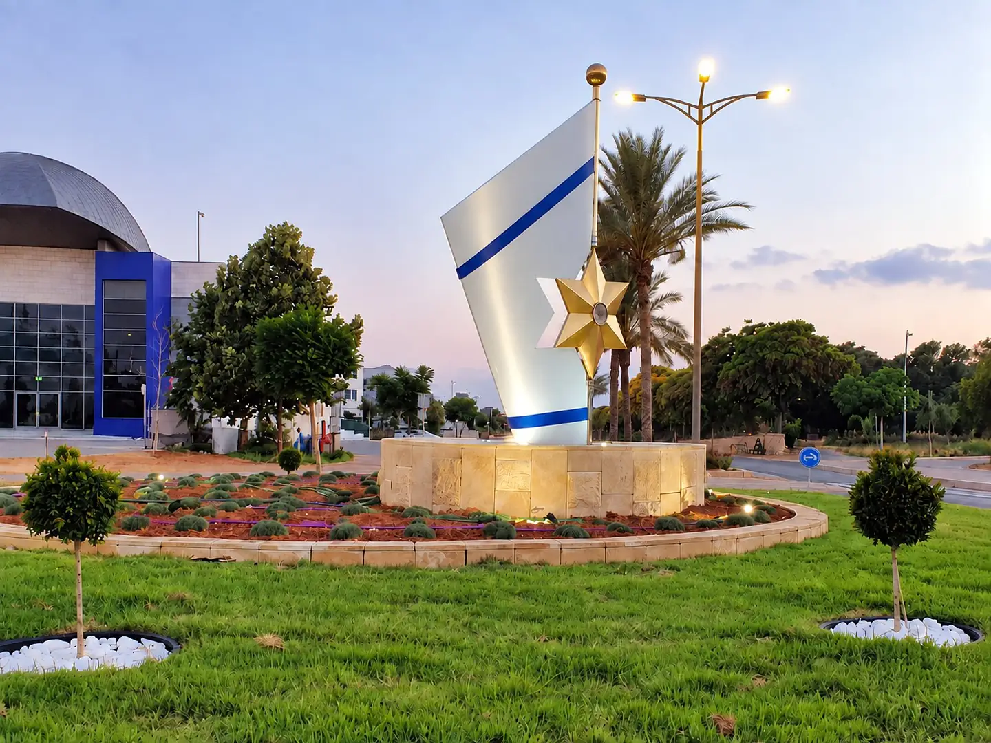 פסל דגל: לוח נירוסטה לבן עם פסים כחולים ומגן דוד זהוב, על בסיס אבן בכיכר, בשעת בין ערביים