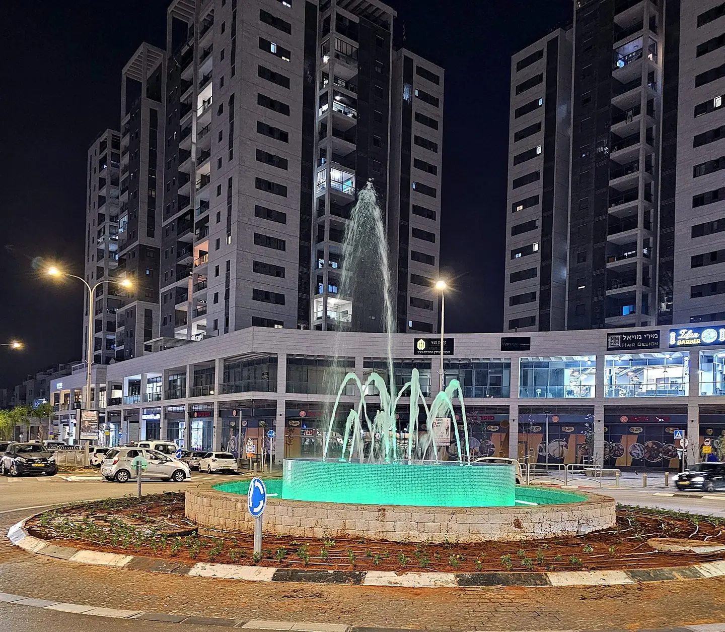 מזרקה בכיכר עירונית בלילה, סילונים מוארים בירוק מול מגדלי מגורים