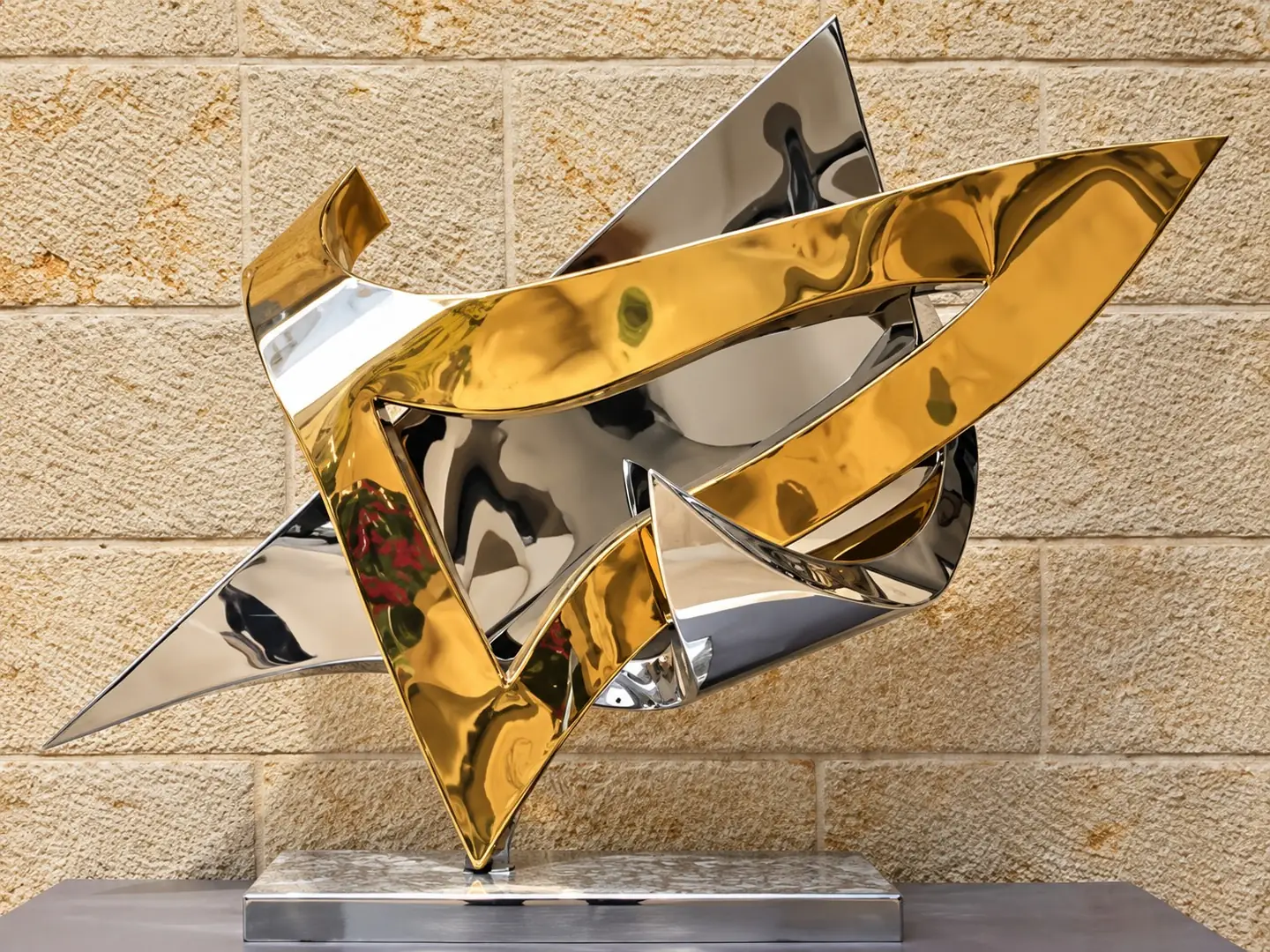 פסל מופשט של משטחי נירוסטה בליטוש מראה ורצועות בגימור זהב, על בסיס נירוסטה, מול קיר אבן